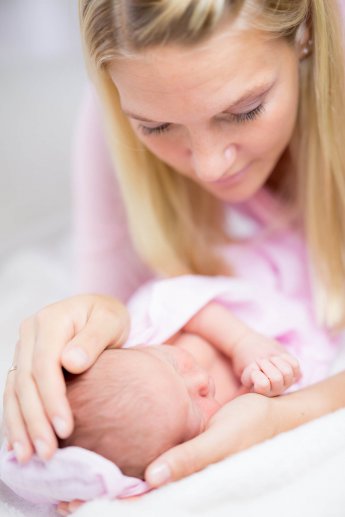 Mutter hält den Kopf des Neugeborenen zwischen den Händen und beugt sich über das Baby. Überwiegend weiß und pink dominieren das süße Bild
