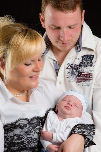 Junges Elternpaar mit Neugeborenem vor schwarzem Hintergrund fotografiert