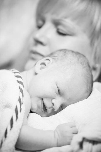 Schwarzweiss-Foto vom lächelnden Baby, das bei der schlafenden Mama im Arm liegt