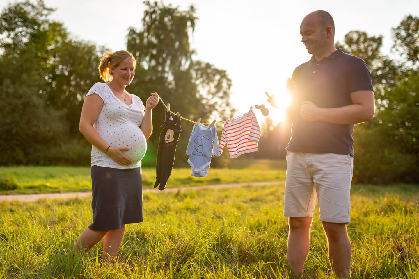 Werdende Eltern halten beim Babybauchshooting eine Wäscheleine mit Babykleidung im Gegenlicht