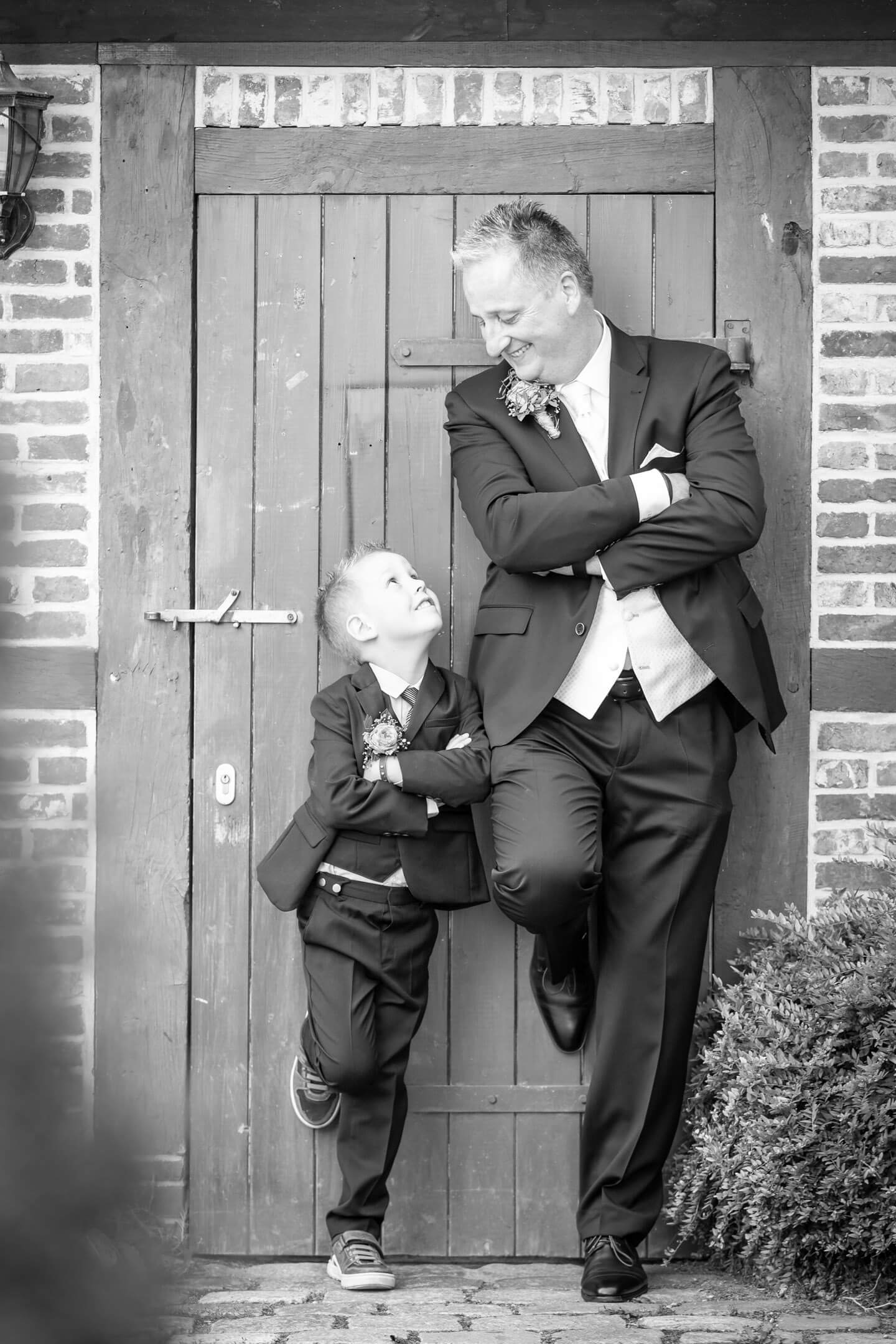 Tolles Hochzeitsfoto: Vater und Sohn lehnen sich in ihren Hochzeitsanzuegen gegen eine Wand und blicken sich an.