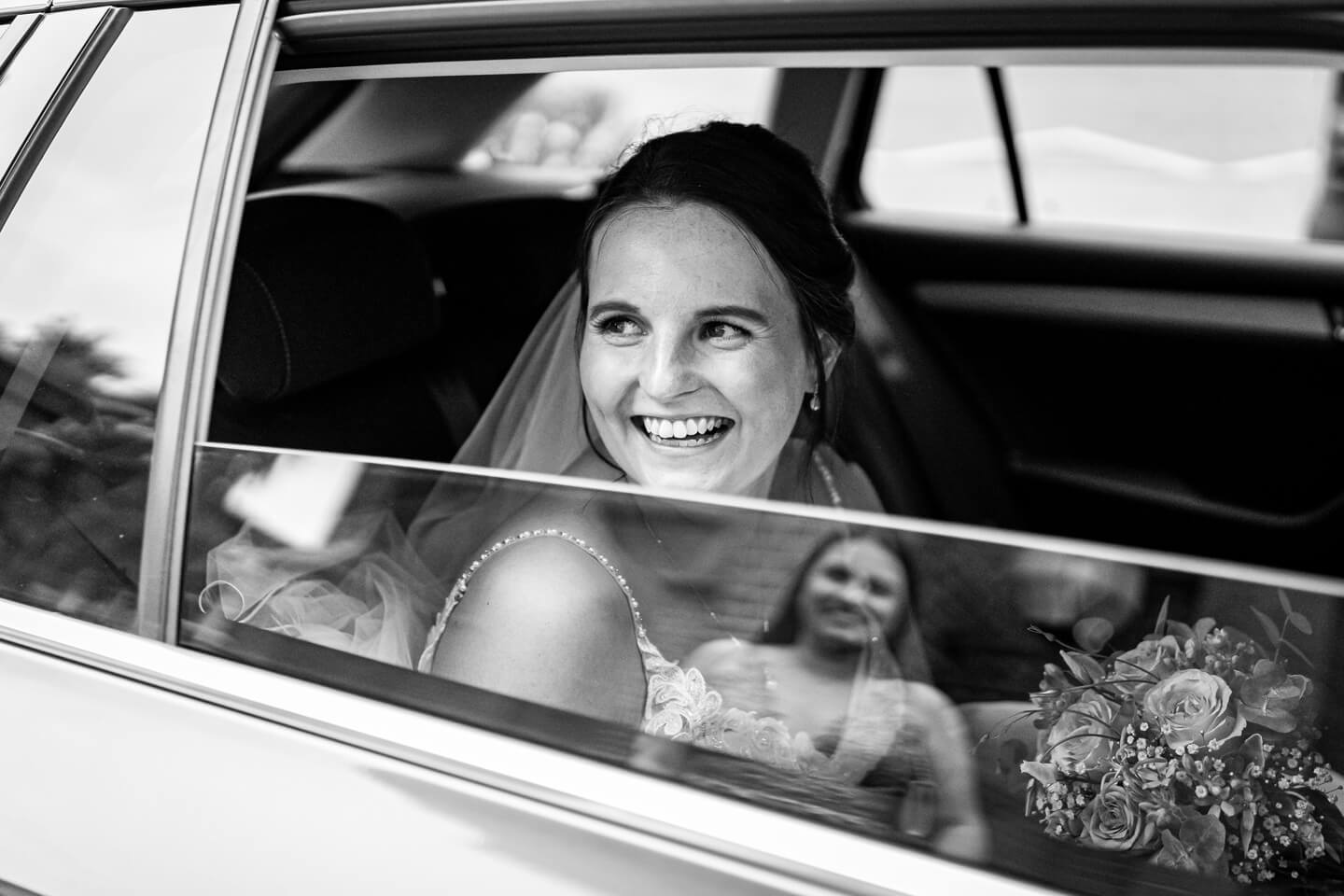 Braut sitzt im Hochzeitsauto, die Schwester spiegelt sich in der Scheibe